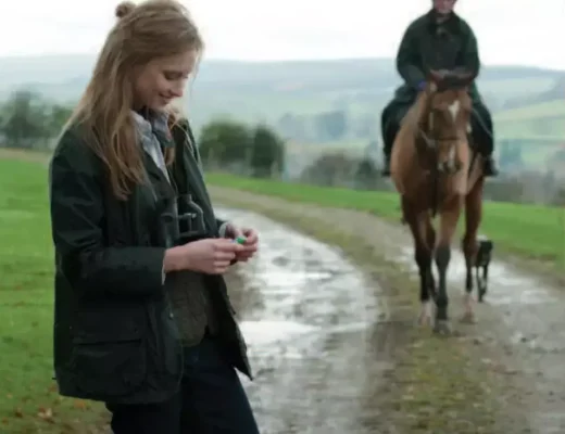 Frau mit dunkelgrüner Barbour-Beadnell-Jacke in britischer Landschaft mit Reiter im Hintergrund