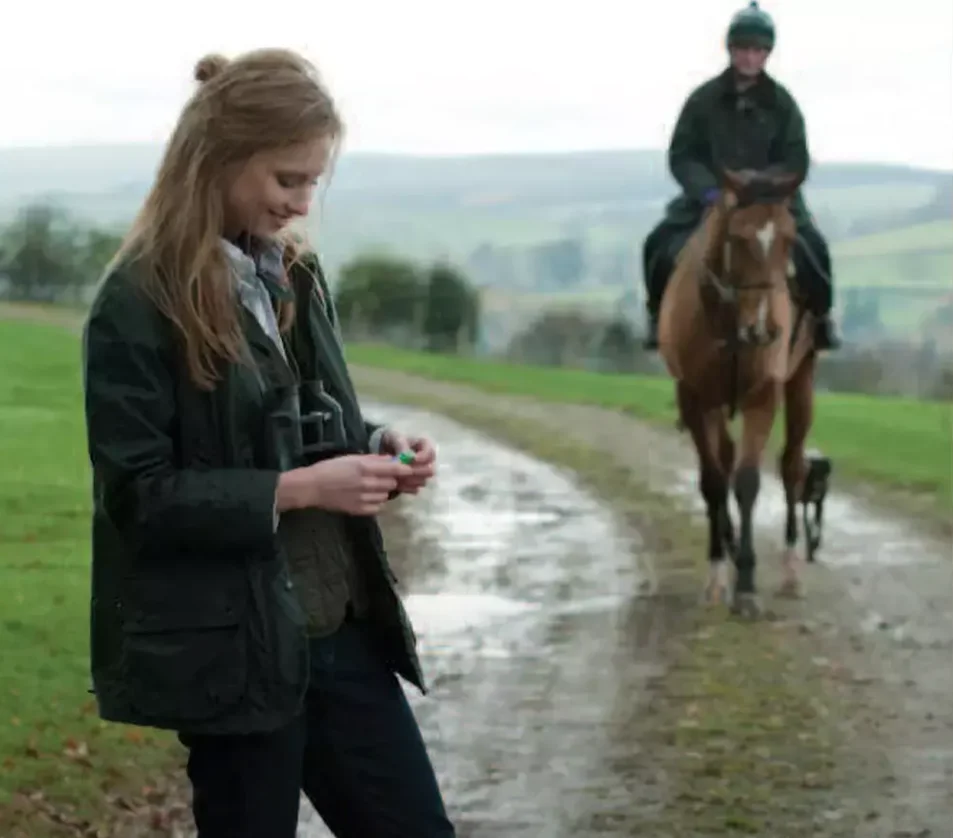 Frau mit dunkelgrüner Barbour-Beadnell-Jacke in britischer Landschaft mit Reiter im Hintergrund