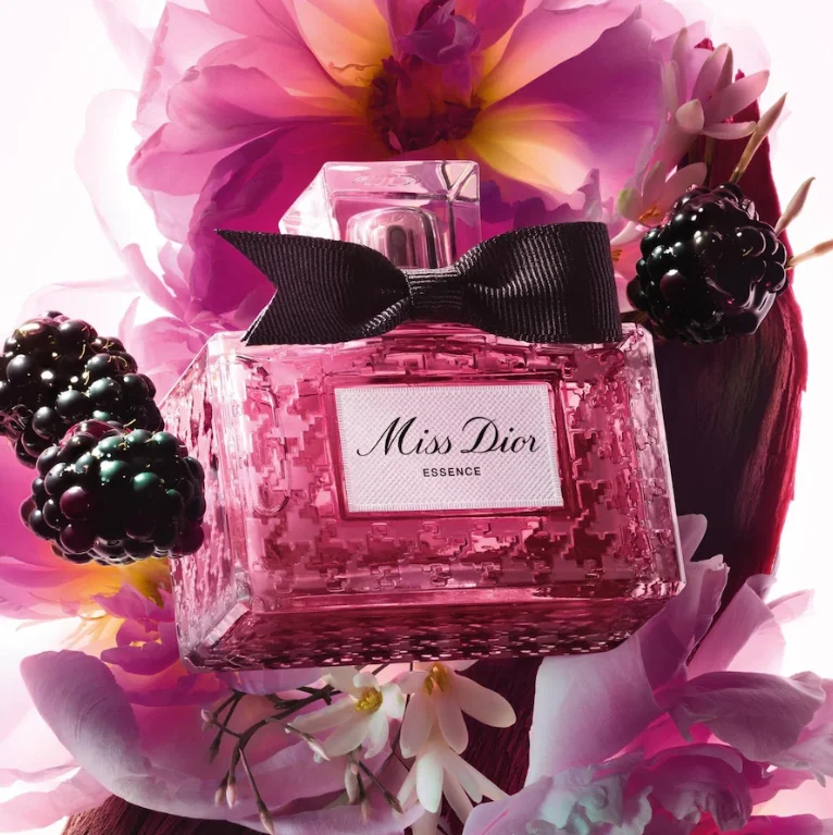Miss Dior Essence Parfum im pinkfarbenen Dior-Flakon mit schwarzer Schleife, umgeben von Brombeeren und Blüten.