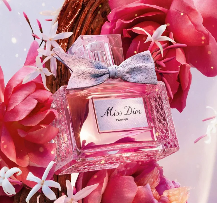 Miss Dior Parfum im rosafarbenen Glasflakon mit silberner Schleife, umgeben von pinkfarbenen Blüten.